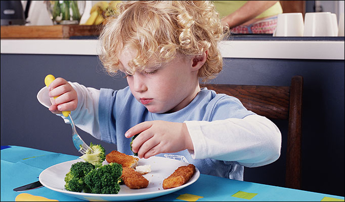 Boy (2-4) eating fish fingers and broccoli, mother at kitchen sink